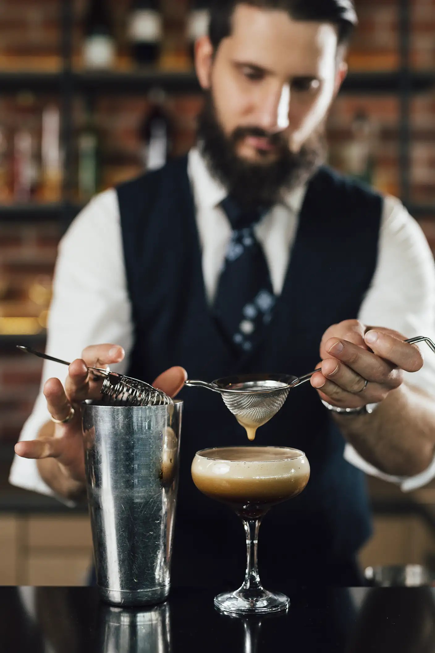 A bearded bartender straining and pouring an espresso martini into a glass
