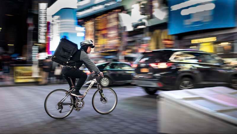 A fast moving photo of a food delivery worker on a bicycle going through a busy city street at night