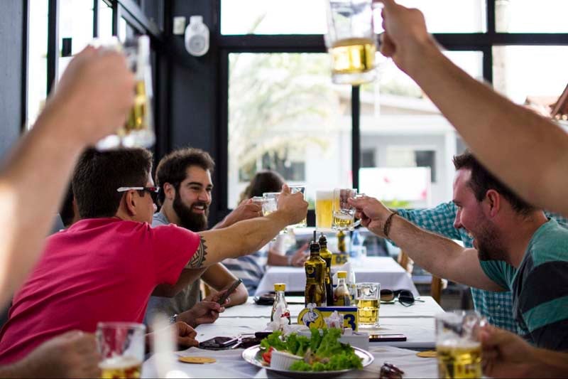 Group of friends sitting at dining table at a bar or brewery toasing and raising beer glasses during happy hour