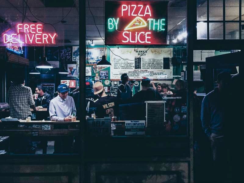 A nightime photo of a busy pizza restaurant offering food delivery services with neon sign