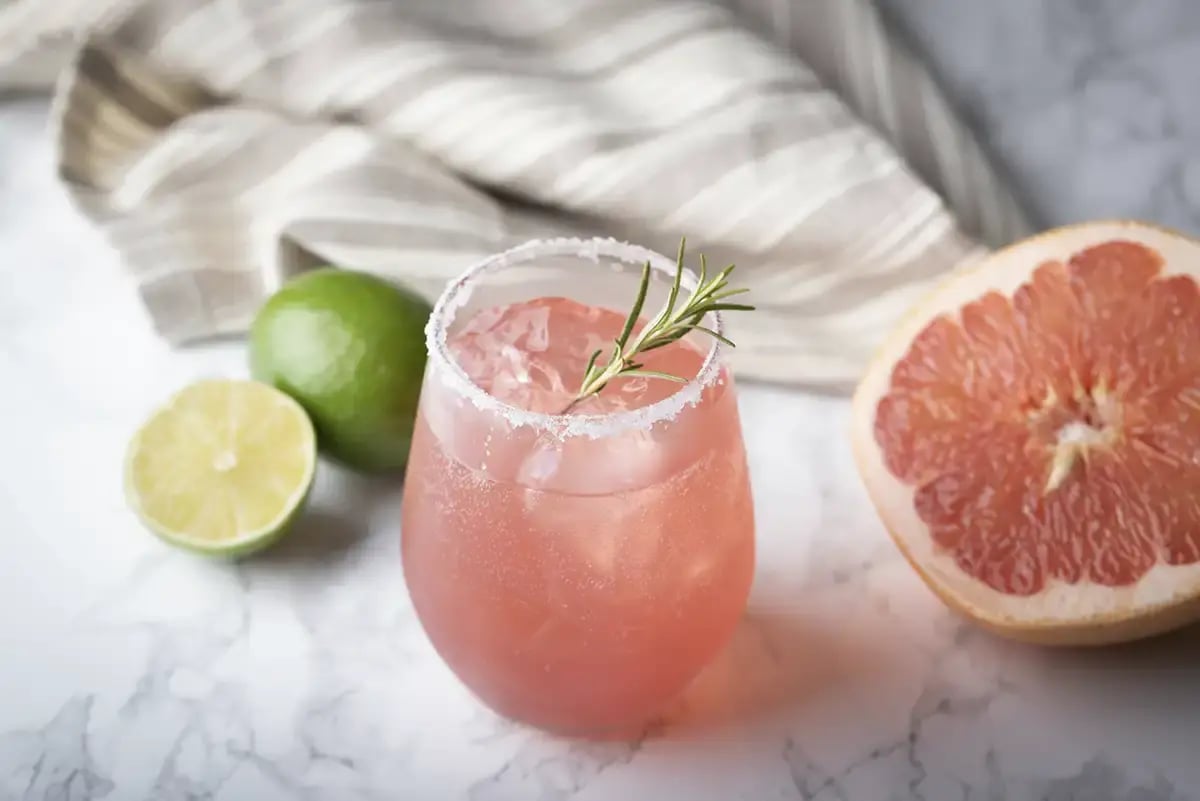 A Paloma tequila cocktail on a marble table with a grapefruit and lime used as accents
