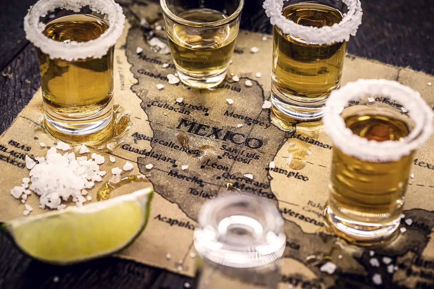 Various shots of tequila and mezcal with limes on top of a canvas map of Mexico